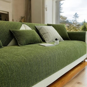 May include: A sofa covered in a textured green fabric with a herringbone pattern. The sofa features a white base, several green rectangular throw pillows, and one square throw pillow with a white and green striped pattern. A small glass bottle sits on the seat.