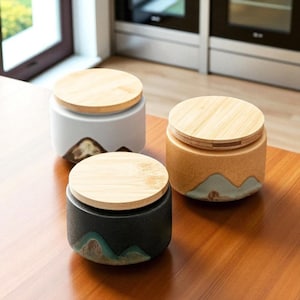 May include: Three ceramic jars with wooden lids sit on a wooden surface. The jars are cylindrical with a mountain design in shades of brown, teal, and white. The lids are made of light-colored wood. The jars are likely for kitchen storage.