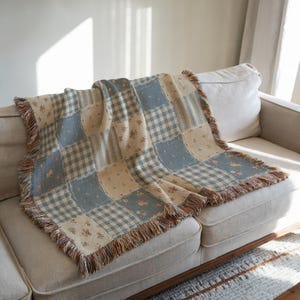 May include: A patchwork throw blanket with a mix of patterns, including gingham, floral, and striped designs in blue, beige, and cream. The blanket has a fringed edge and is draped over a beige sofa.