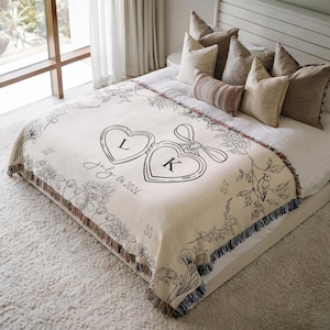 May include: Cream-colored throw blanket with black floral and heart-shaped design. Features the letters "L" and "K" inside hearts, with "July 04 2026" below. The blanket has a fringed edge and is placed on a bed.