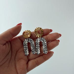 Puede incluir: Un par de pendientes con una parte superior circular dorada y una parte inferior en forma de U blanca con lunares negros. Los pendientes se sostienen en una mano sobre un fondo blanco. Un accesorio único.
