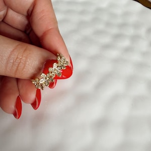 Puede incluir: Un anillo dorado con un diseño floral con pequeñas piedras preciosas transparentes. El anillo se sostiene sobre un fondo blanco. El diseño floral del anillo está formado por múltiples flores pequeñas.