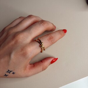 Puede incluir: Un anillo dorado con diseño de nudo se lleva en un dedo. El anillo es el punto focal, con un estilo simple y elegante. La mano tiene esmalte de uñas rojo y un pequeño tatuaje de cuatro estrellas.