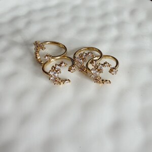 Puede incluir: Cuatro anillos dorados con piedras preciosas transparentes y brillantes dispuestas en racimos. Los anillos se exhiben sobre una superficie blanca texturizada con un borde dorado. Los anillos parecen ser de metal.