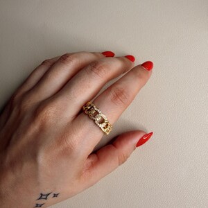 Puede incluir: Un anillo dorado con eslabones de cadena y pequeñas piedras transparentes engastadas en el centro. El anillo está en un dedo con esmalte de uñas rojo. El fondo es de color beige neutro.