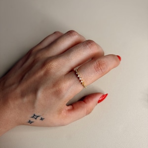 Puede incluir: Un delicado anillo de oro con pequeñas piedras brillantes se lleva en un dedo. El anillo se encuentra sobre un fondo neutro. Las uñas de la persona están pintadas de rojo y un pequeño tatuaje de cuatro estrellas es visible en la mano.