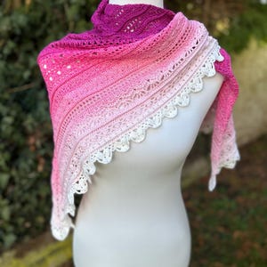 Peut inclure: Un châle en crochet rose et blanc fait à la main, présenté sur un mannequin. Le châle présente un dégradé de nuances de rose, passant du magenta profond à une bordure en dentelle blanche douce. Le travail de crochet complexe est visible.