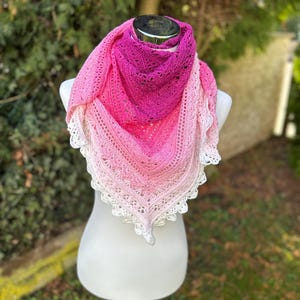 Könnte beinhalten: Ein handgehäkelter Schal in Rosatönen, der von einem tiefen Magenta oben zu einer weißen Spitzentrimmung übergeht. Der Schal wird auf einer weißen Schaufensterpuppe präsentiert, die das detaillierte Stichmuster und das zarte Design hervorhebt.