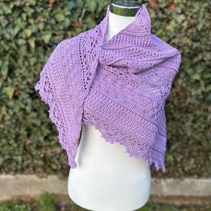 Könnte beinhalten: Ein lavendelfarbener Häkelschal, der über einer weißen Schaufensterpuppe drapiert ist. Der Schal hat komplizierte Lochmuster an den Rändern und am Körper. Der handgefertigte Schal wird vor einem Hintergrund aus grünem Laubwerk präsentiert.