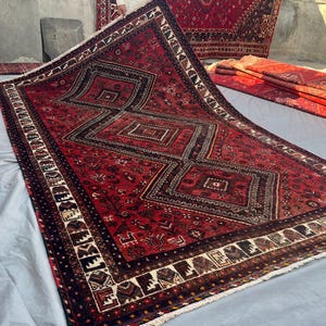 Puede incluir: Una alfombra rectangular grande con un fondo rojo intenso y tres medallones centrales en forma de diamante. La alfombra presenta intrincados patrones geométricos en negro, blanco y marrón, enmarcada por un borde ancho y detallado.
