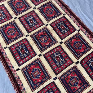 Puede incluir: Una alfombra rectangular con un patrón repetitivo de cuadrados rojos y azul marino sobre un fondo crema. Cada cuadrado presenta un diseño central y está enmarcado por un borde azul marino. Los bordes de la alfombra tienen un borde detallado.