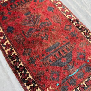 May include: A rectangular, hand-woven rug with a deep red field and intricate geometric patterns in black and brown. The border features a repeating floral design in black, beige, and red. The rug's texture appears soft and plush.