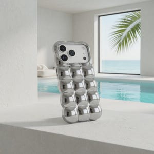 May include: A silver phone case with a unique, textured design. The case features a series of rounded, square-shaped bumps across its surface. The phone case is shown against a backdrop of a pool and ocean view.