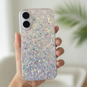 May include: Clear phone case with a glittery, iridescent design. The case has a clear border and a dual-lens camera cutout. The glitter is a mix of silver, blue, and purple, creating a shimmering effect. The phone case is held in a hand.