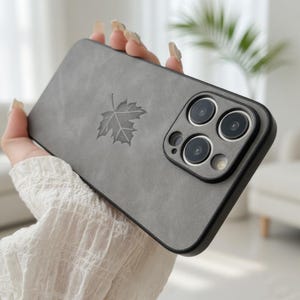 May include: A gray phone case with a leaf design. The case has a matte finish and a black border. The phone case has three camera lenses.