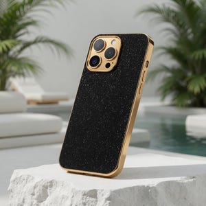 Puede incluir: Funda de teléfono negra con purpurina y borde dorado. La funda tiene forma rectangular con esquinas redondeadas y un recorte para la cámara. La funda del teléfono está en ángulo, mostrando su diseño y características.