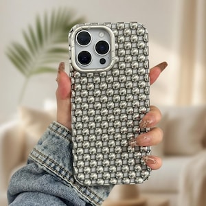 Könnte beinhalten: Silberne Handyhülle mit einem strukturierten, perlenbesetzten Design. Die Hülle hat ein metallisches Finish und einen Kameraausschnitt mit drei Linsen. Die Handyhülle wird in einer Hand gehalten.