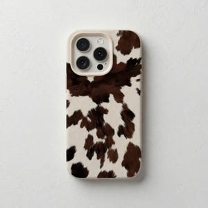 Könnte beinhalten: Eine Handyhülle mit Kuhfellmuster. Die Hülle hat einen cremefarbenen Rand und ein braun-weiß geflecktes Design. Die Kameralinsen sind oben sichtbar.