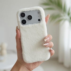 May include: A white, fluffy phone case with a soft, fur-like texture. The case has a rectangular shape with cutouts for the phone's camera and buttons. The case is held in a hand.