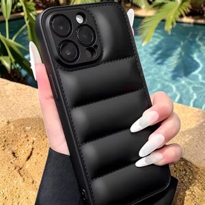 Puede incluir: Funda de teléfono negra acolchada con un recorte para la lente de la cámara. La funda tiene un diseño acolchado y cosido y un acabado liso y mate. La funda del teléfono se muestra sobre un fondo de piscina y palmeras.