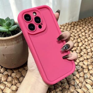 Pode incluir: Uma capa de telefone rosa brilhante com um módulo de câmera elevado. A capa tem um acabamento liso e uma borda contrastante. A capa do telefone é exibida em um fundo tecido, com uma pequena planta suculenta em vaso visível.