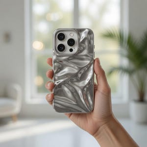 May include: Silver phone case with a rippled, metallic texture. The case has a square shape with rounded corners and a camera module with three lenses. The phone case is held in a hand against a blurred background.