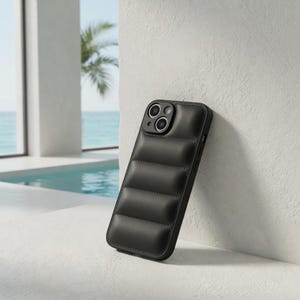Puede incluir: Funda de teléfono negra con diseño acolchado. La funda está apoyada contra una pared blanca, con una piscina y el mar de fondo. La funda tiene un recorte para la cámara.