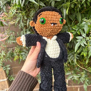 May include: A handmade crochet doll featuring a brown face, black hair, and bright green eyes. The doll wears a black suit jacket over a white shirt, with small white fangs visible below the nose. The plush figure has a soft, textured yarn finish.