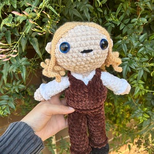 May include: A handmade crochet doll featuring blonde yarn hair, large blue plastic eyes, and a small mouth with two white fangs. The doll wears a white long-sleeved shirt, a dark brown velvet-textured vest, matching brown trousers, and black shoes.