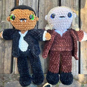 May include: Two handmade crochet dolls made of soft plush yarn. The left doll has brown skin, black hair, green eyes, and a black suit with a white shirt. The right doll has light skin, blonde hair, blue eyes, and a brown suit with a white shirt.