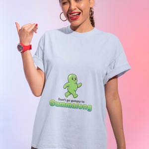 Könnte beinhalten: Ein hellblaues kurzärmeliges T-Shirt mit einer Grafik eines kleinen grünen Gummibärchen-Charakters. Unter der Figur steht der Text "Don't go gummy to GUMMALONG" in grüner und schwarzer Schrift. Das T-Shirt hat einen Rundhalsausschnitt und umgekrempelte Ärmel.