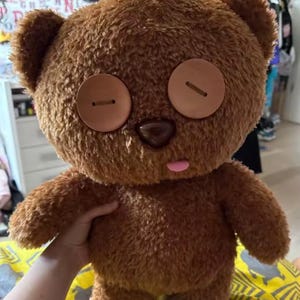 May include: A brown plush teddy bear with large button eyes and a pink tongue sticking out. The bear has a round head, small ears, and a brown nose. The bear is soft and fluffy, with a friendly expression.