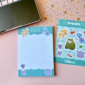 May include: A laptop sits on a pink surface next to a notepad and sticker sheet. The notepad has lined pages and illustrations. The sticker sheet features various cartoon characters and the text "Pokopia" and "Telltoons."