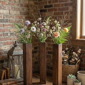 Può includere: Tre vasi alti di legno scuro, ognuno con una diversa composizione di fiori freschi e verde. I vasi rettangolari sono su un pavimento di legno davanti a un muro di mattoni. Una lanterna rustica e dei libri sono sullo sfondo.