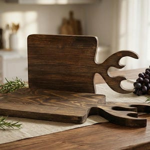 Puede incluir: Tablas de cortar de madera marrón oscuro con asas únicas en forma de cuerno. Las tablas se exhiben sobre una mesa de madera con un camino de mesa de lino, ramitas de romero, aceitunas, uvas y queso.