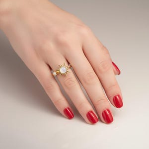 Könnte beinhalten: Ein goldfarbener Ring mit einem Sonnenmuster und einem zentralen, runden, weißen Stein. Der Ring hat auch einen kleinen, runden, klaren Stein an der Seite. Die Hand trägt roten Nagellack.