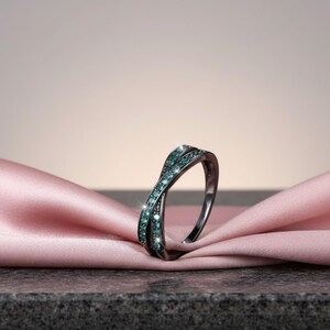 May include: A black-toned ring with a crisscross design, adorned with teal-colored gemstones. The ring is displayed on a pink satin fabric, showcasing its elegant and modern style. The gemstones sparkle, adding a touch of glamour.