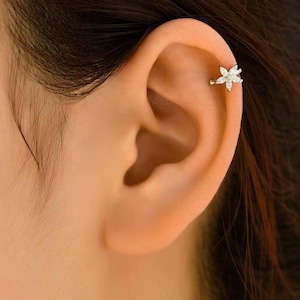 Puede incluir: Primer plano de una oreja con un pendiente hélix de color dorado. El pendiente tiene forma de flor con múltiples piedras claras y brillantes. La oreja es de tono claro y el pelo es castaño oscuro.