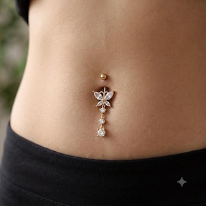 Könnte beinhalten: Ein goldfarbenes Bauchnabelpiercing mit einem schmetterlingsförmigen Anhänger, der mit klaren, marquise-geschliffenen Kristallen besetzt ist. Das Design umfasst eine hängende Kette mit drei runden, klaren Kristallen unter dem Schmetterling.