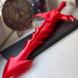 Puede incluir: Una vibrante espada decorativa roja con una hoja y empuñadura dentadas y detalladas, apoyada sobre una base rectangular negra. El diseño de la espada presenta bordes afilados y una punta, evocando un tema de fantasía o medieval.