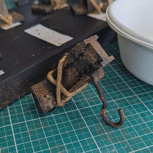 May include: A weathered, rusty metal hook with a coiled spring and a square, riveted body. A length of tan rope is wrapped around the body. The hook is set against a green grid surface, suggesting a crafting or workshop setting.