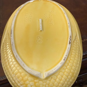 Pode incluir: Uma travessa de servir vintage em cerâmica amarela em forma de espiga de milho. A travessa tem uma superfície texturizada que imita grãos de milho e um interior liso. As palavras "Shawnee USA" e "Oven Proof" são visíveis. O número "68" também está presente.