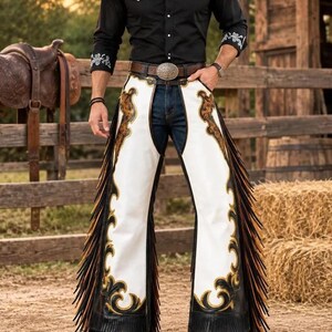 May include: A black long-sleeved button-down shirt with white floral embroidery on the shoulders and cuffs, paired with white leather chaps featuring black and brown scrollwork patterns, long fringe along the sides, and dark blue denim jeans.