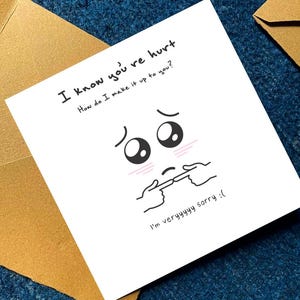 May include: A white greeting card with the text "I know you're hurt. How do I make it up to you?" and "I'm veryyyyy sorry :(". The card features a cartoon face with sad eyes and hands pointing to the mouth. The card is placed on a blue surface with a brown envelope.