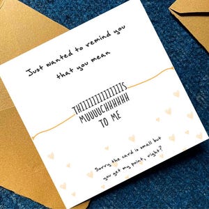 Puede incluir: Una tarjeta blanca con el texto "Just wanted to remind you that you mean THIIIIIIIIIIIIIIS MUUUUCHHHHHH TO ME". La tarjeta est&aacute; decorada con peque&ntilde;as formas de coraz&oacute;n y est&aacute; colocada sobre una superficie azul con un sobre dorado.