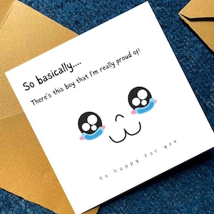 May include: A white greeting card with a cartoon face and the text "So basically.... There's this boy that I'm really proud of! So happy for you." The card is placed on a dark blue surface, next to a gold envelope.