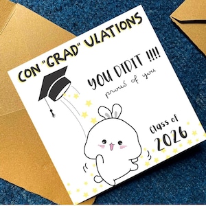 K&ouml;nnte beinhalten: Abschlusskarte mit dem Text "CON 'GRAD' ULATIONS" und "YOU DID IT!!!! proud of you". Die Karte zeigt eine Cartoon-Abschlusskappe und einen wei&szlig;en Hasen-Charakter mit den Worten "Class of 2026". Die Karte ist wei&szlig; mit gelben Sternen.