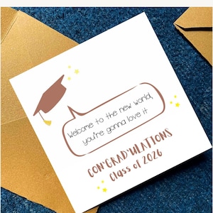 May include: A graduation greeting card with a brown mortarboard illustration and text that reads, "Welcome to the new world, you're gonna love it." The card also says "CONGRATULATIONS Class of 2026" with yellow star accents. The card is in a gold envelope.