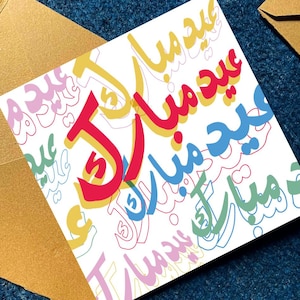 Peut inclure: Une carte de v&oelig;ux blanche avec une calligraphie arabe color&eacute;e disant "Eid Mubarak" en rouge, jaune, bleu et vert. La carte est pos&eacute;e sur une surface bleu fonc&eacute; avec une enveloppe dor&eacute;e.