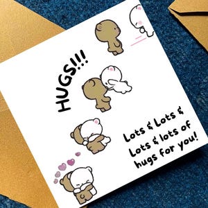 May include: A white greeting card with the words "HUGS!!!" and "Lots & Lots & Lots & lots of hugs for you!" in black text. The card features cartoon bears hugging, with pink hearts. The card is placed on a blue surface.